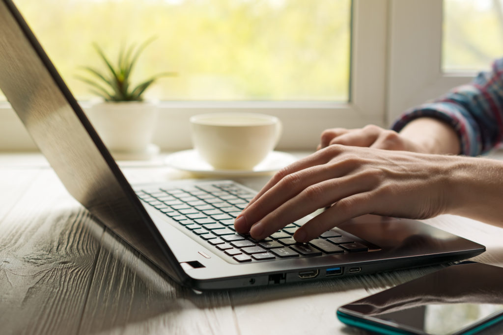 Hands typing on a keyboard. Modern work place at home with black notebook, smartphone and cup of coffee on light window background